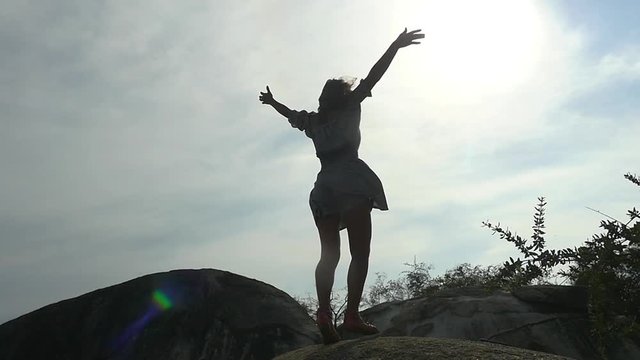 Silhouette of young joyful woman standing on the rock rises her hands against the sky through the sun. slow motion. 1920x1080