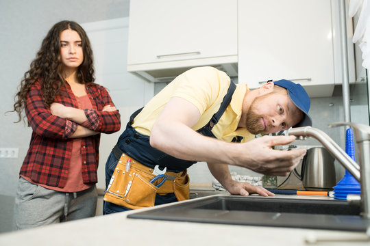 Serious concentrated young bearded plumber in cap standing at kitchen counter and checking dropping faucet while house owner with crossed arms standing behind him