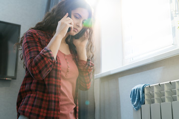 Serious sad young woman with curly hair calling repair service while having leaking radiator in flat