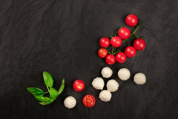 Mozzarella cheese balls with fresh basil leaves and cherry tomatoes, the ingredients of the Italian Caprese salad, shot from the top on a black background with copy space