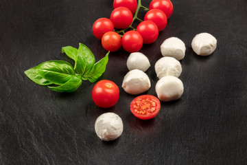 Mozzarella cheese balls with fresh basil leaves and cherry tomatoes, the ingredients of the Italian Caprese salad, on a black background with copy space