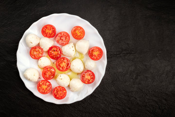 An overhead photo of a plate of Italian Caprese salad with Mozzarella cheese and cherry tomatoes, shot on a dark background with copy space