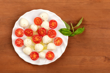 An overhead photo of a plate of Italian Caprese salad with Mozzarella cheese, cherry tomatoes, and basil leaves, shot on a dark rustic wooden background with copy space