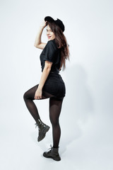 Fototapeta na wymiar Happy stylish dark-haired girl dressed in a black t-shirt, shorts, tights and cap is posing on the white background in the studio