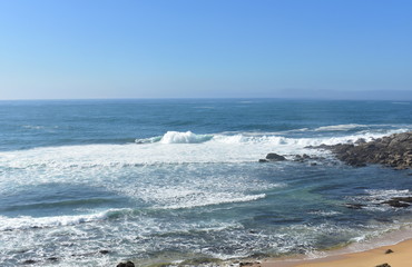 Wild beach with rocks and big waves with foam and mist. Blue sky, sunny day, Galicia, Spain.