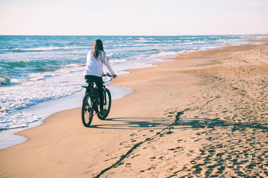 Woman rides a Bicycle along the sea on a sandy beach. Back view. - Powered by Adobe