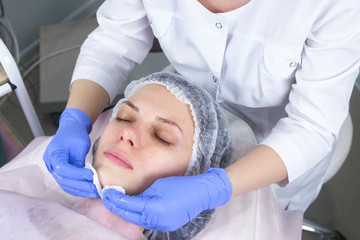 Naklejka premium Cosmetologist wipes the face of a young woman with cotton pads. Hands of the beautician in gloves and a face closeup. Beauty treatments for the face.