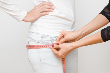 Woman's hands measuring young, slim girl's hips with pink measuring tape. Part of body on gray...