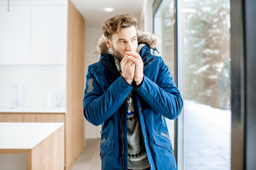 Man dressed in winter clothes feeling cold standing near the window at home with no heating