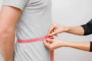Woman's hands measuring young, slim man's waist with pink measuring tape. Part of body on gray...