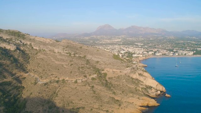 Aerial cinematic scenic view, flying slow above blue sea along big yellow rocky mountain coastline, yachts and city view at sunset sunrise golden hour time. Beautiful summer autumn view and soft light