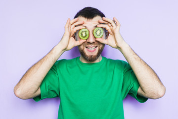 Young bearded man holding slices of green kiwi fruit in front of his eyes, smiling and surprised. Light purple background, copy space.