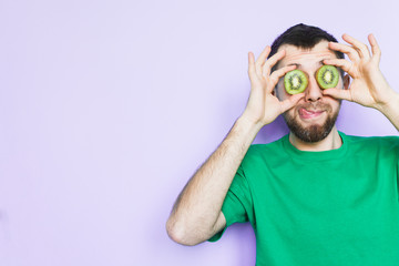 Young bearded man holding slices of green kiwi fruit in front of his eyes, showing tongue. Light purple background, copy space.