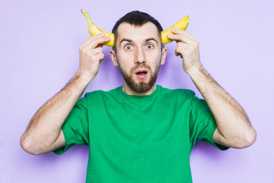 Young Bearded Man Holding Cut In Half Yellow Banana On The Level Of Temples, Surprised And Shocked Face Expression. Light Purple Background, Copy Space.