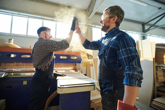 Positive satisfied bearded factory workers standing at workbench with particleboard and making dusty high five while greeting each other at workplace
