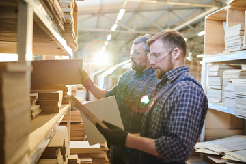 Serious brutal workers in safety goggles standing at shelves full of wooden details and choosing...