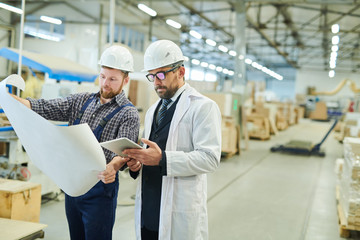 Serious engineers in hardhats working with project blueprint: concentrated young worker showing sketch to businessman in white coat using digital tablet at factory workshop