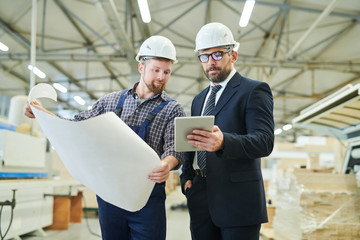 Serious handsome middle-aged investor in eyeglasses using digital tablet and showing online data to engineer with blueprint
