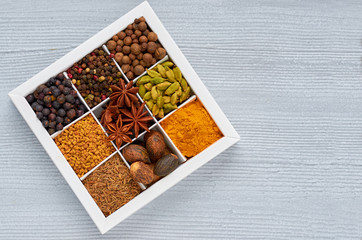 Spices and herbs in the box on the gray kitchen table: anise stars, fragrant pepper, cinnamon, nutmeg, bay leaves, paprika close up - ingredients for tasty food and cuisine. Top view with copy space