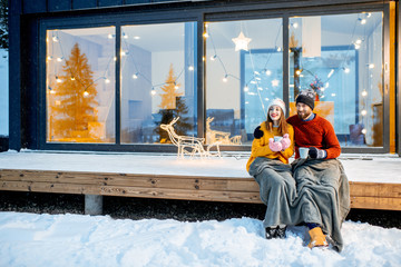 Young couple warming up with plaid and hot drinks sitting on the terrace of the modern house in the...