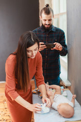 The father makes a photo on the phone as his wife changes a diaper to their child