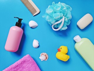 Pink packaging of liquid soap, green shampoo bottle, sponge, cotton towel, comb, shells and yellow rubber duckling. Flat lay baby cosmetics for skin and hair care