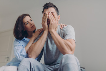 Photo of depressed male dressed casually, his beautiful woman comforts him, sit together in...