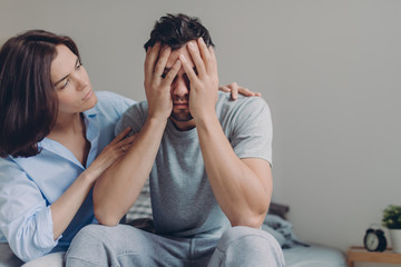 Stressful young man cries from despair, loving mother tries to calm him, pose together at bed...