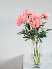 Beautiful pink Living Coral peony bouquet in glass vase on table with white linen tablecloth. Vertical. Copy space for text. Toned filter