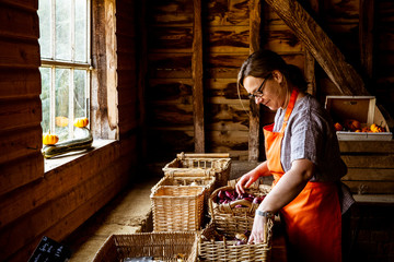 Woman wearing apron standing in farm shop, arranging baskets with vegetables.