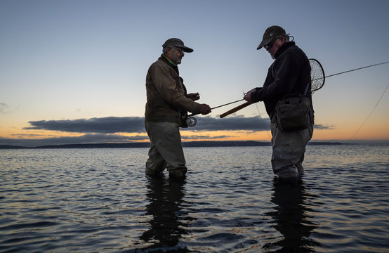 A male fly fisherman watches his guide work putting on a new fly to try for salmon or trout on  a salt water beach at Fort Flagler State Park in northwest Washington State, USA