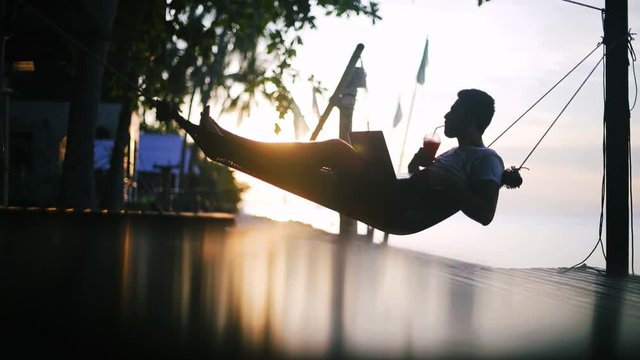 A young, handsome man working on a laptop lying in a hammock with a cocktail in his hands at sunset. Freelancer sipping a cocktail and working on travel.