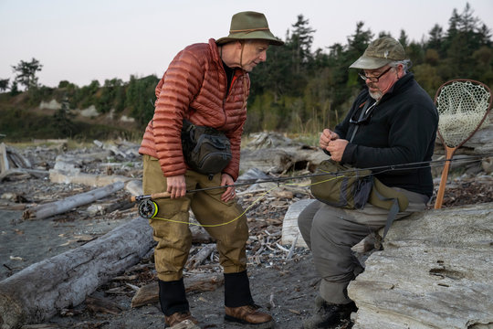 A male fly fisherman watches his guide work putting on a new fly to try for salmon or trout at a salt water beach.