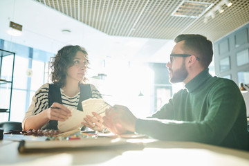 Obraz premium Portrait of two modern designers discussing business sitting at table in office lit by sunlight