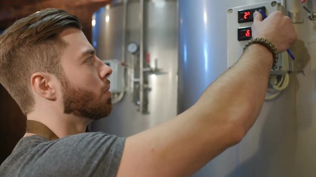 a young handsome bearded brewer man on a background of beer tanks and a brewery making notes in a tablet
