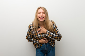 Blonde young girl over white wall smiling a lot while putting hands on chest