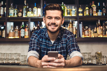 Young bartender leaning on bar counter holding smartphone smiling cheerful