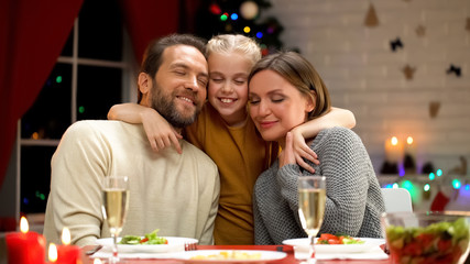Happy family having Xmas dinner, daughter hugging parents, decorations twinkling