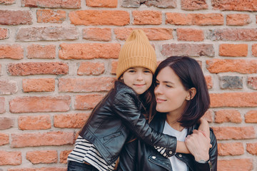 Fototapeta na wymiar Affectionate mother with dark hair recieves warm hug from daughter, wear leather jackets, pose together against brick wall. Fashionable little girl in hat embraces her mum. Family and children concept