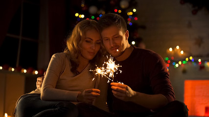 Loving couple with Bengal lights sitting near Xmas tree, making wish in New Year