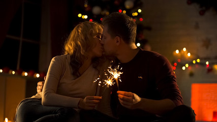 Young couple with Bengal lights kissing tenderly near sparkling Christmas tree