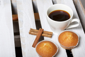 A cup of freshly brewed coffee and muffins. Nearby are cinnamon sticks. Located on a wooden box.