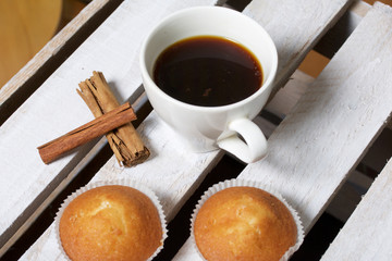 A cup of freshly brewed coffee and muffins. Nearby are cinnamon sticks. Located on a wooden box.