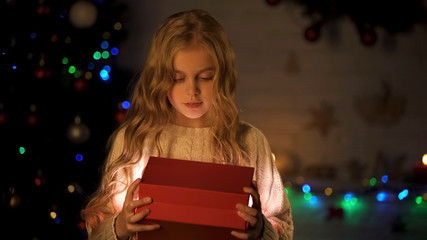 Adorable girl opening gift box near Christmas tree, childish dream come true