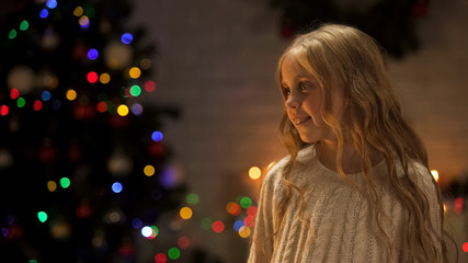 Little girl looking out Santa near glowing Christmas tree, holiday anticipation