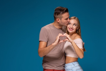 Naklejka premium Happy couple dressed casual clothes, making heart shape from fingers, studio portrait on blue background