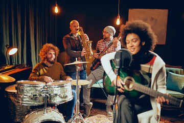 Mixed race woman singing and playing guitar while sitting on chair with legs crossed. In background drummer, saxophonist and bass guitarist.