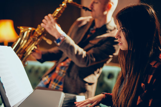 Young Caucasian woman with long brown hair playing electric piano and in background saxophonist playing his instrument. Home studio interior.