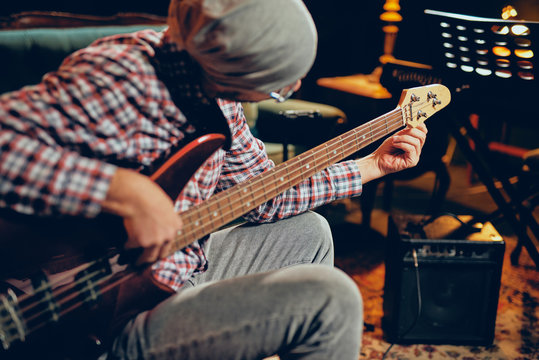 Young Caucasian man with hat on head playing bass guitar while sitting in home studio.