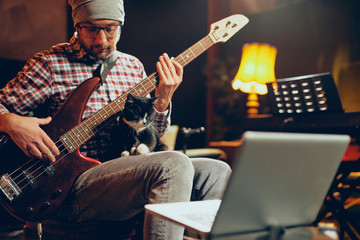 Young Caucasian man with hat on head playing bass guitar while sitting in home studio. - obrazy, fototapety, plakaty